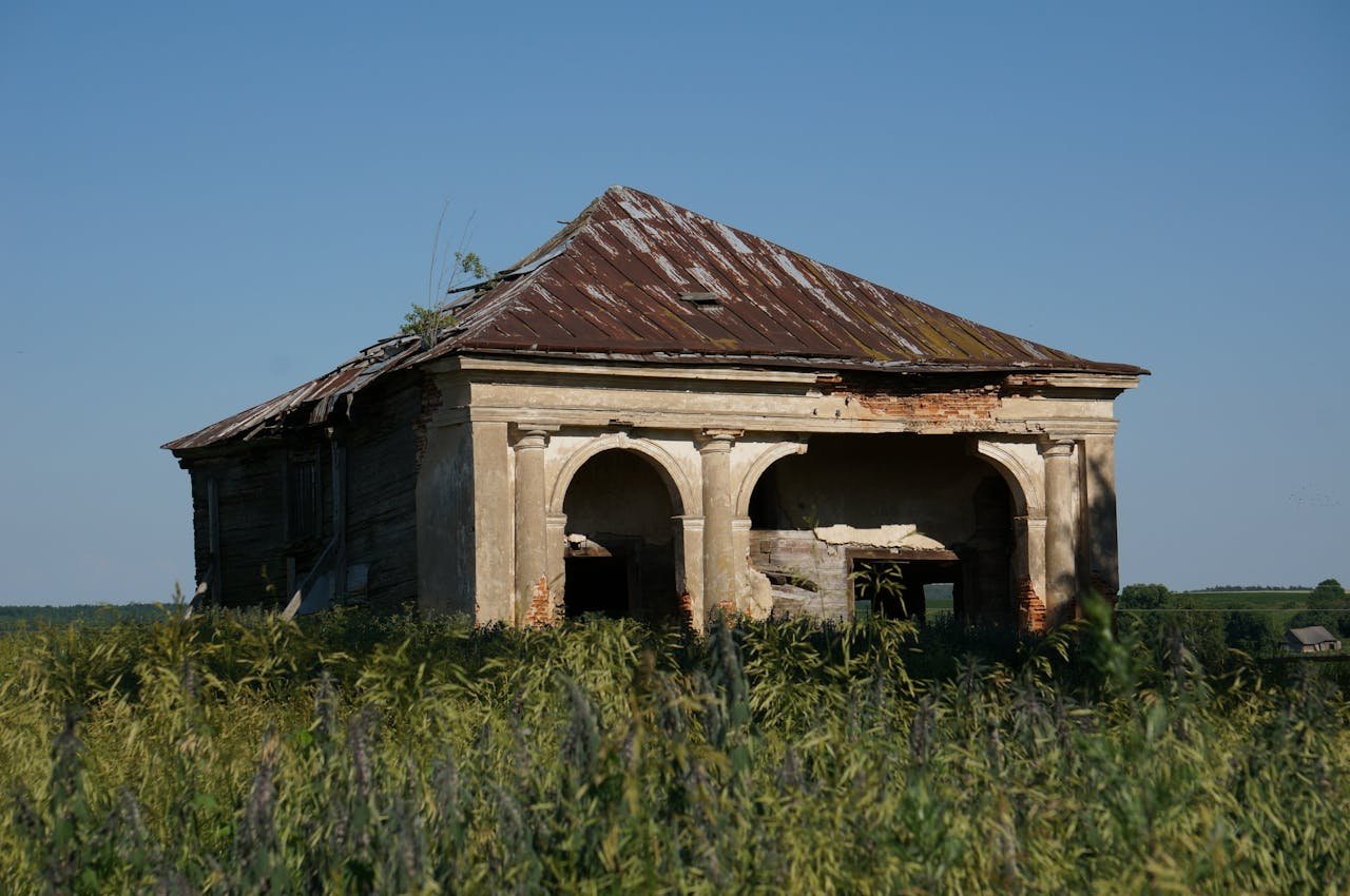 Dilapidated rural house surrounded by lush vegetation under clear sky, showcasing rustic charm.